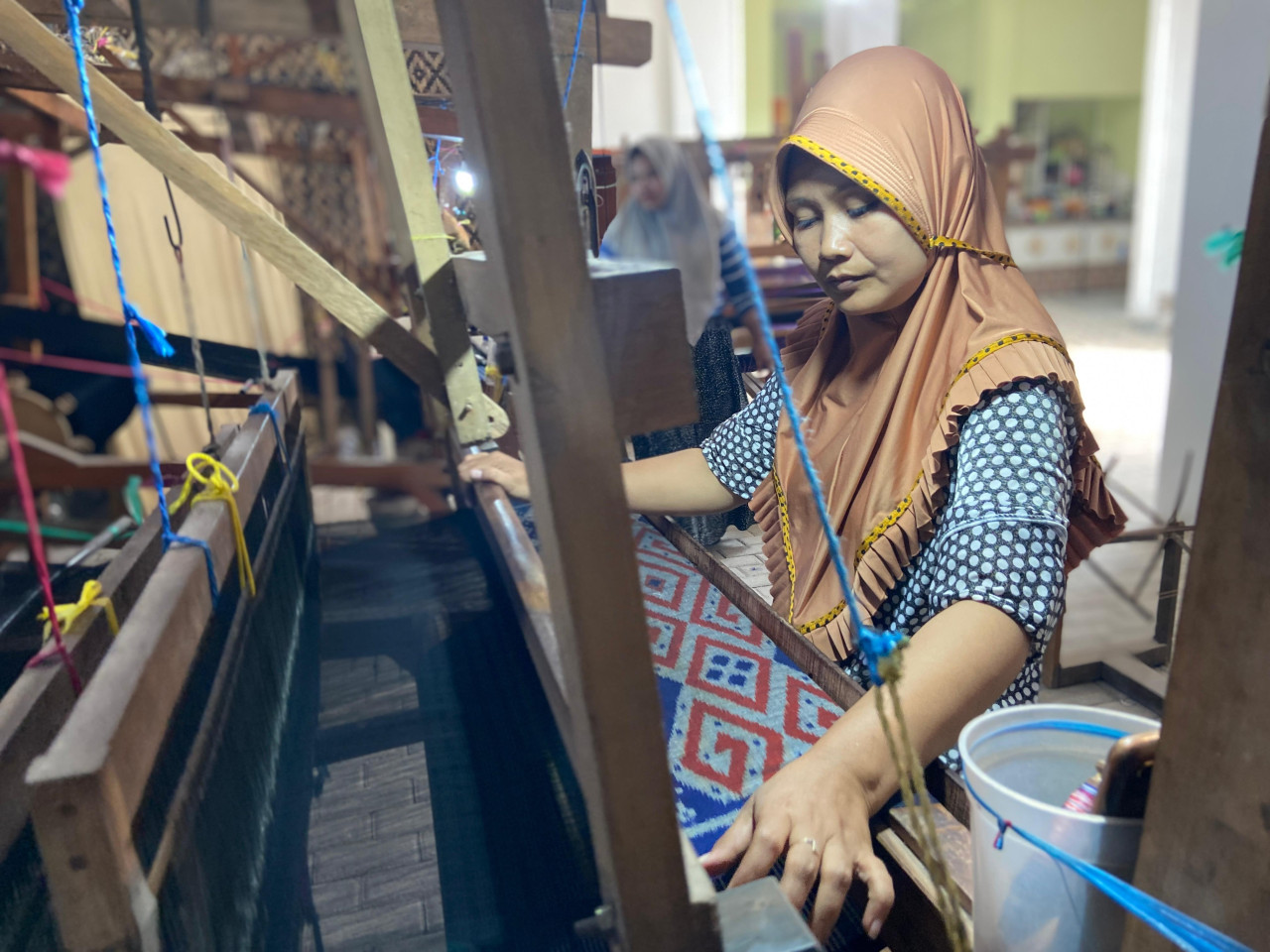 Mira (35) sedang menenun di gerai kedua Medali Emas. Dia sudah bekerja sejak tahun 2000-an. Dia cukup senang bisa membantu ekonomi keluarga. Suaminya bekerja sebagai kuli bangunan.