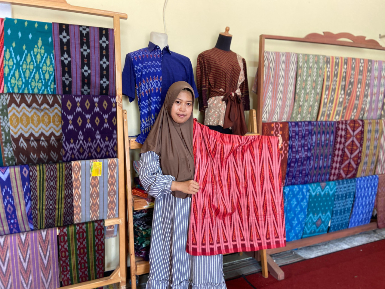 Yusna Qurrota A’yun (32), anak pertama dari Siti Ruqoyah berfoto di depan deretan kain tenun ikat di gerainya yang memesona. Dia memegang kain dengan motif khas Kota Kediri, Salur Tirto, berwarna merah yang dihasilkan dari air panas dengan pembakaran sempurna menggunakan LPG. Terlihat sangat tajam dan menyala.