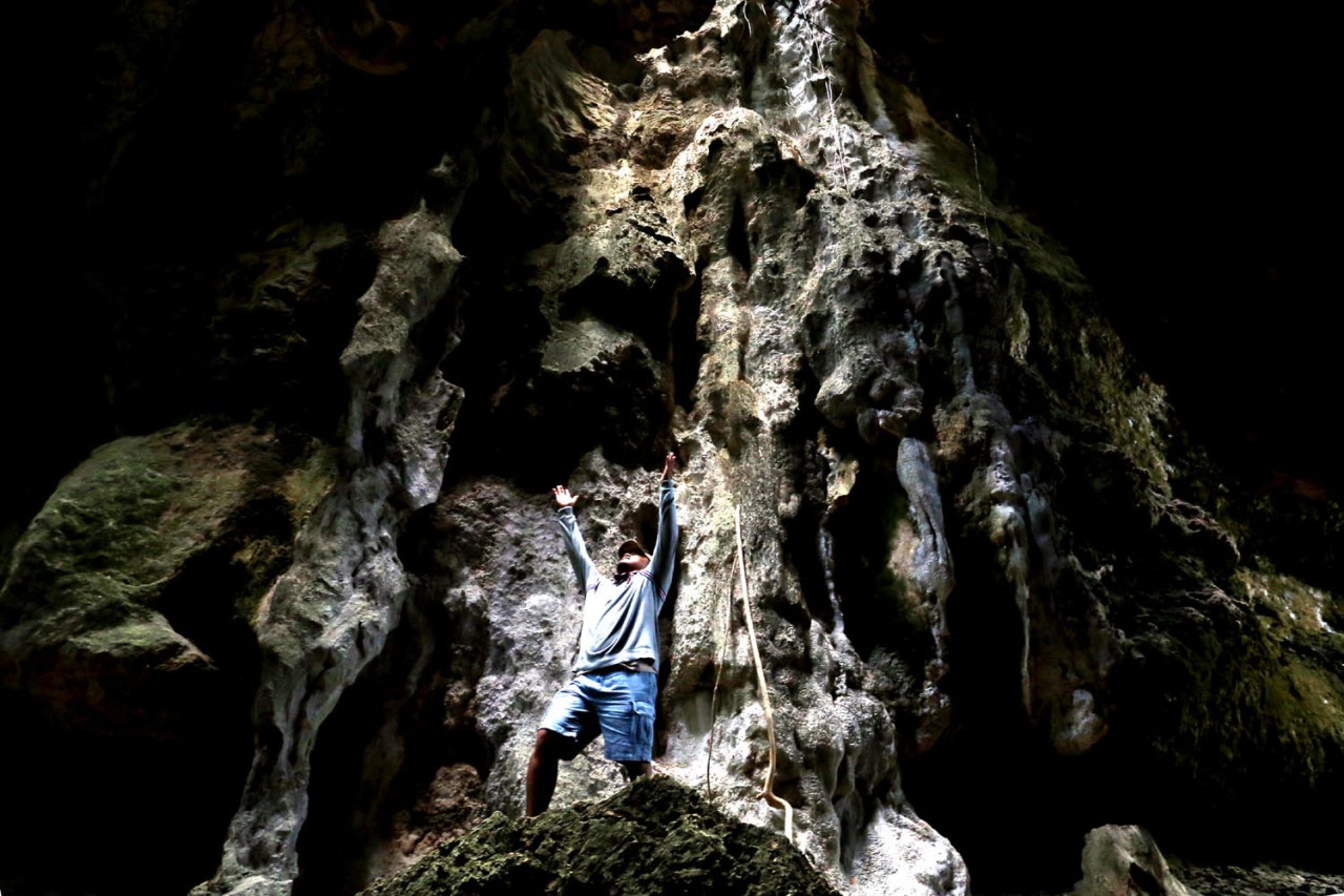 Wisatawan menghadap ke atas dengan tangan menengadah saat berada di dalam  Goa Batu Cermin. Cahaya yang menembus celah bebatuan menciptakan suasana magis. (Foto: Ali Masduki/JatimNow.com)
