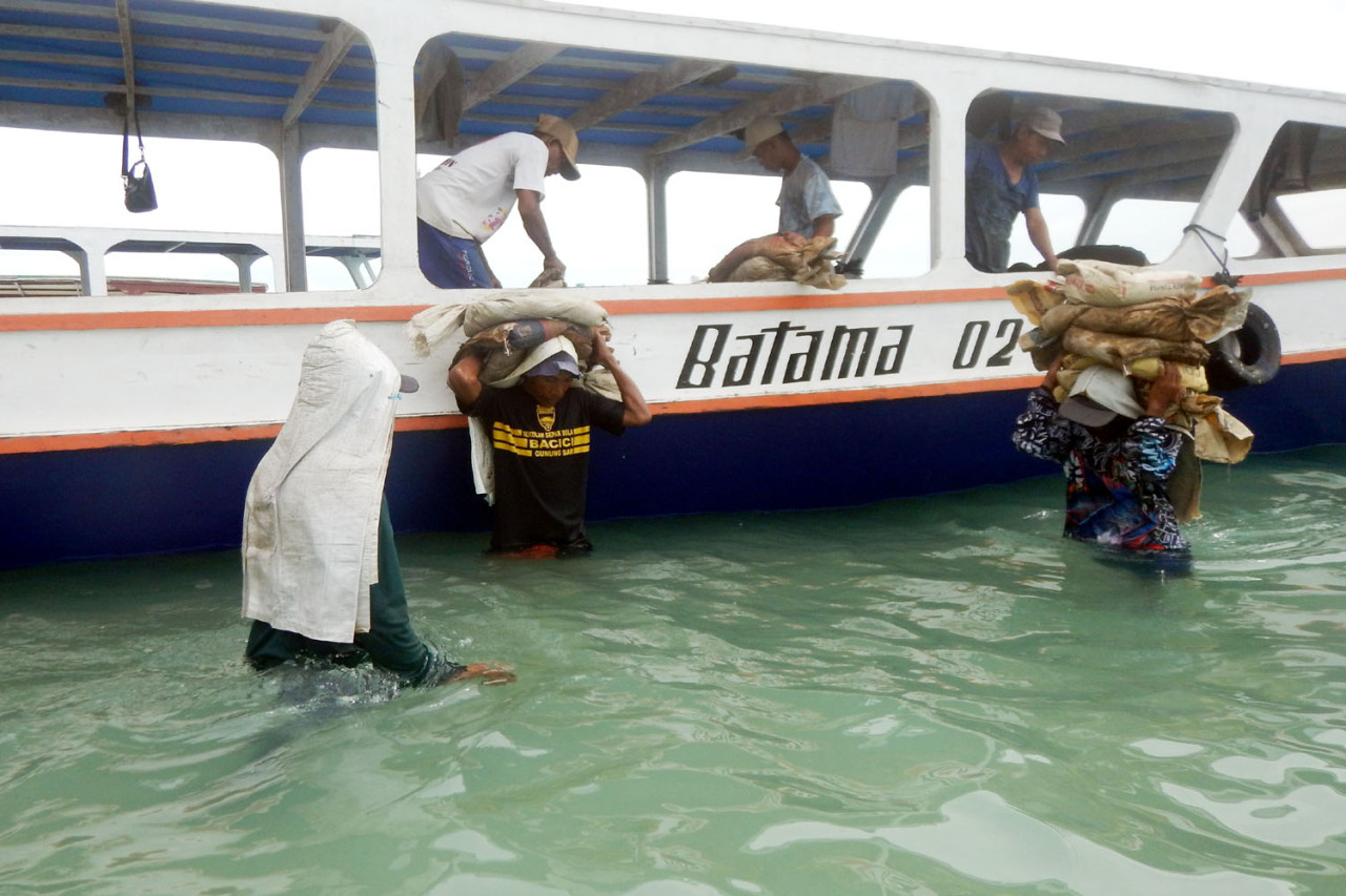 Kuli angkut memindahkan karung material bangunan dari perahu ke daratan Gili Trawangan, Lombok. Tanpa alat berat dan kendaraan bermotor, bongkar muat dilakukan di laut dangkal. Bahu manusia menggantikan mesin, menjadi bagian dari ongkos mahal dan proses panjang di balik pembangunan pulau wisata ini. (Foto: Ali Masduki/jatimnow.com)