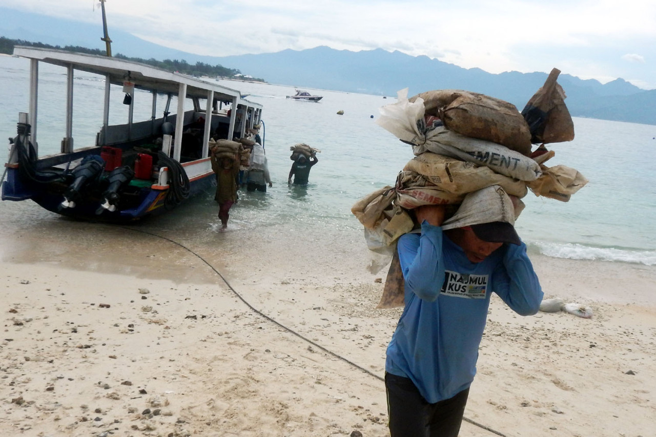 Seorang kuli angkut membawa karung semen dari bibir pantai Gili Trawangan, Lombok. Material bangunan diturunkan dari perahu kecil dan dipanggul melintasi pasir tanpa bantuan alat berat. Di pulau wisata tanpa kendaraan bermotor ini, setiap bangunan berdiri di atas tenaga manusia dan ongkos logistik yang mahal.(Foto: Ali Masduki/jatimnow.com)