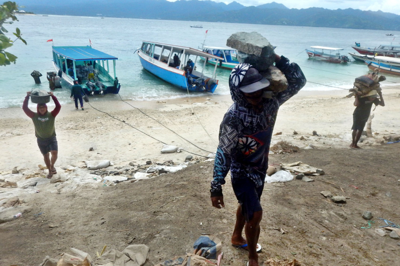 Kuli angkut membawa batu bangunan dari bibir pantai menuju lokasi pembangunan di Gili Trawangan, Lombok. Tanpa kendaraan bermotor dan alat berat, material dipanggul menanjak dari pantai. Di balik indahnya pulau wisata, proses pembangunan bergantung pada tenaga manusia dan ongkos logistik yang tidak murah.(Foto: Ali Masduki/jatimnow.com)