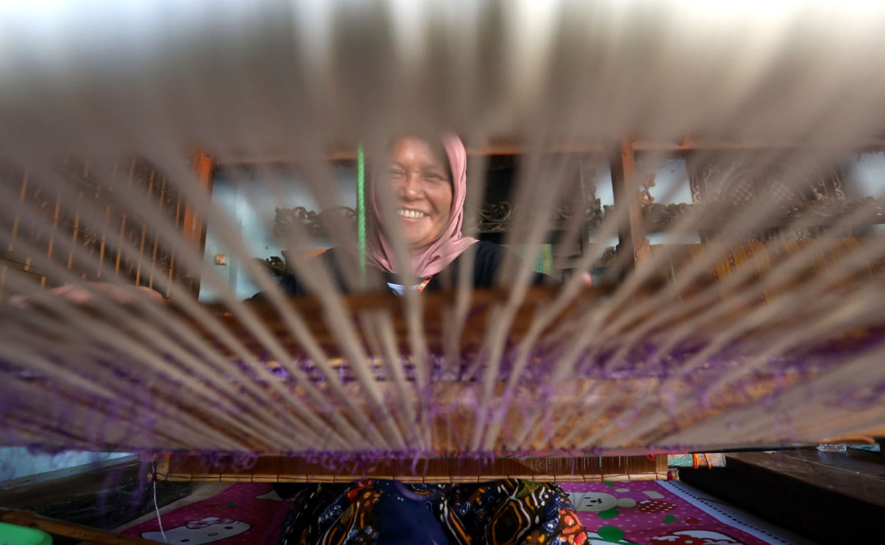 Senyum seorang penenun Desa Sukarara terlihat di balik helaian benang yang bergerak, menggambarkan kebanggaan dan ketekunan perempuan Sasak dalam merawat tradisi tenun songket yang telah diwariskan turun-temurun. (Foto: Ali Masduki/jatimnow.com)