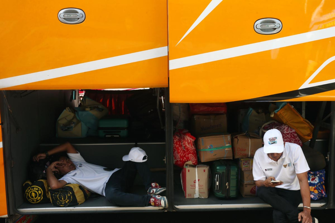 Seorang peserta beristirahat di bagasi bus di tengah tumpukan barang bawaan saat pemberangkatan program mudik gratis di Surabaya, Senin (17/3), sementara peserta lain menunggu sambil memeriksa ponsel. Program yang digelar PTPN I Regional 5 ini diminati masyarakat sebagai alternatif perjalanan aman dan hemat menjelang Lebaran. (Foto: Ali Masduki/jatimnow.com)