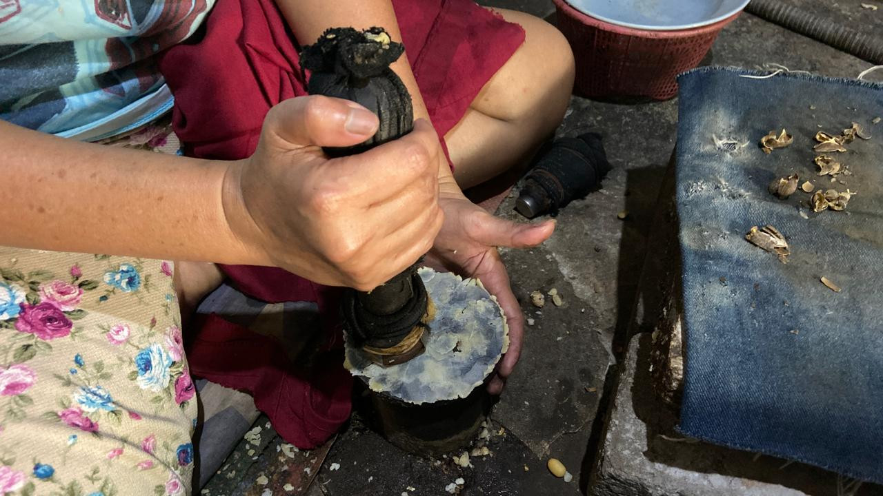 Ida Kristiani (49) menumbuk biji melinjo yang telah disangrai hingga menjadi kepingan tipis di dapur rumahnya di Koplakan, Kecamatan Pare, Kabupaten Kediri, Kamis (8/10/2026). Proses pemipihan membutuhkan ketepatan dan keterampilan tangan agar menghasilkan bentuk emping yang sesuai. Cedera pada jari-jarinya menjadi pengalaman menyakitkan sekaligus lucu bagi Ida saat pertama kali menggeluti pekerjaan ini ketika masih duduk di bangku SMA. (Foto: Yanuar Dedy/jatimnow.com)